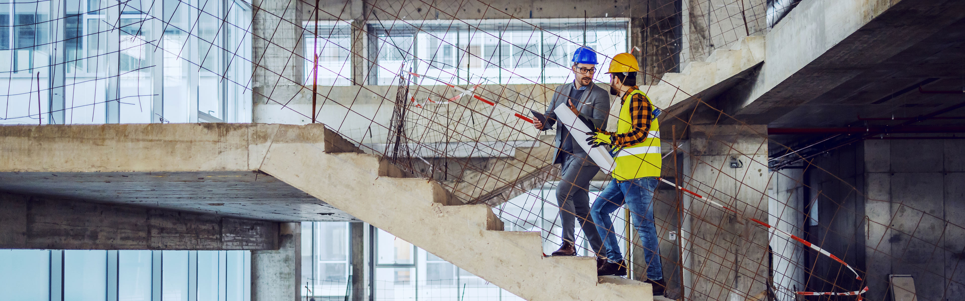 Zwei Bauleiter auf einer Baustelle in einem unfertigen Gebäude, die auf einer Beton-Treppe stehen und Baupläne sowie Details besprechen, beide mit Schutzhelmen und reflektierender Sicherheitskleidung ausgestattet.