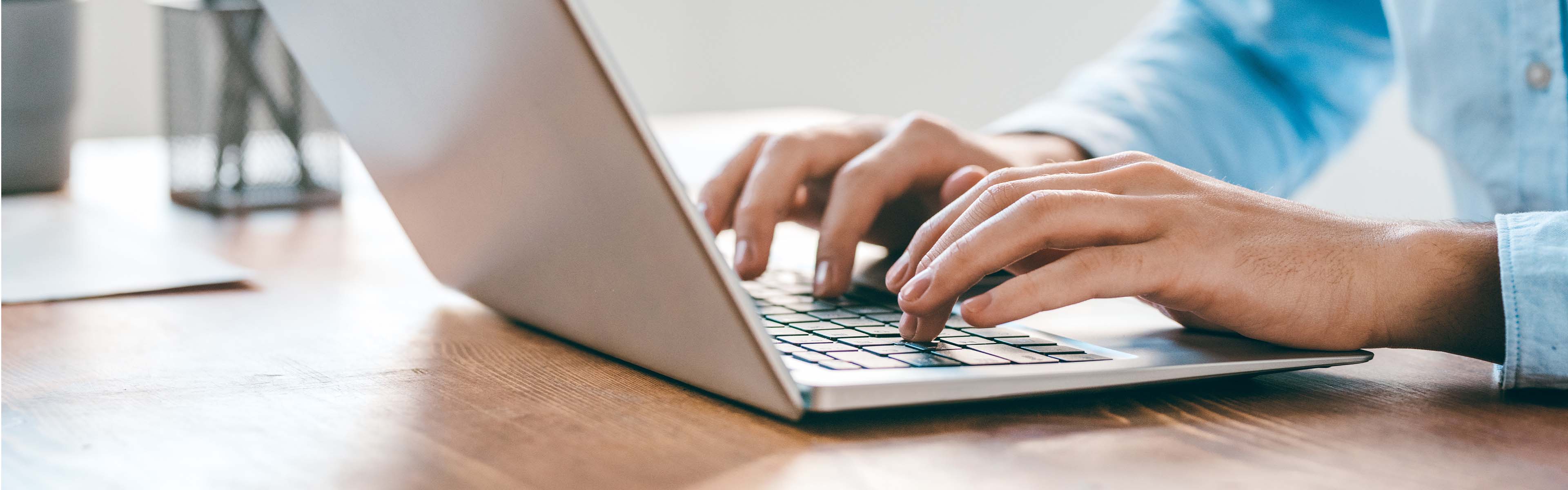 Nahe Ansicht von Händen, die auf der Tastatur eines Laptops tippen, auf einem hölzernen Schreibtisch in einem gut beleuchteten Arbeitsumfeld.