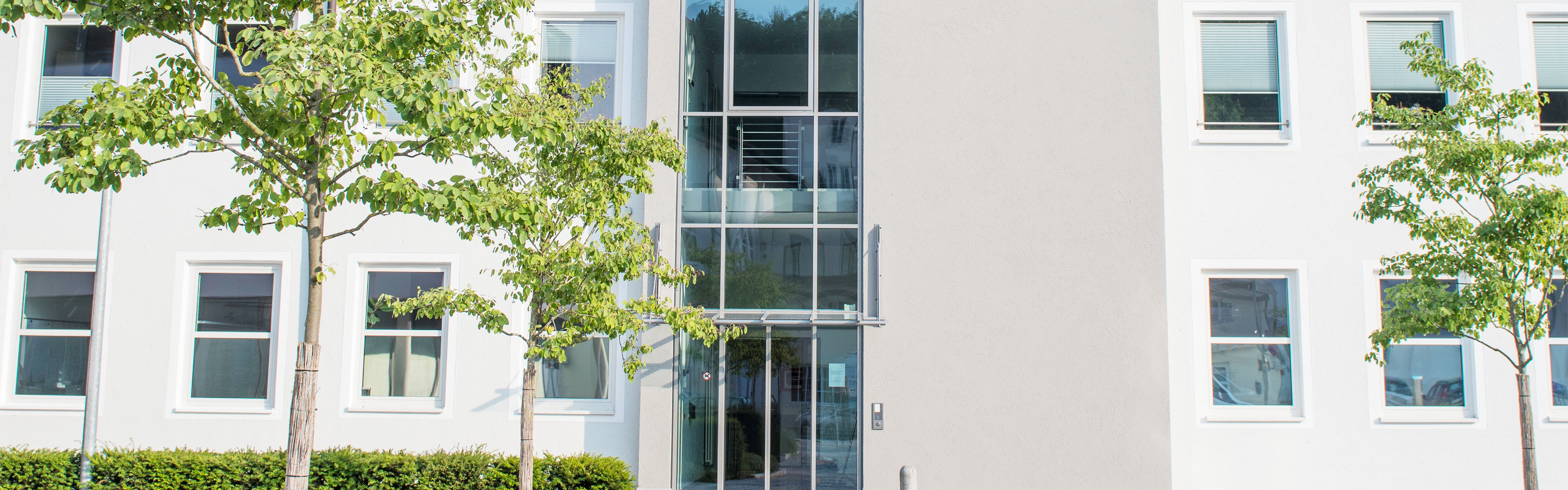 Gebäude der Dachau CityCom mit großen Glasfenstern und weißen Wänden, umgeben von gepflegten Bäumen und grünen Hecken, bei strahlendem Tageslicht.