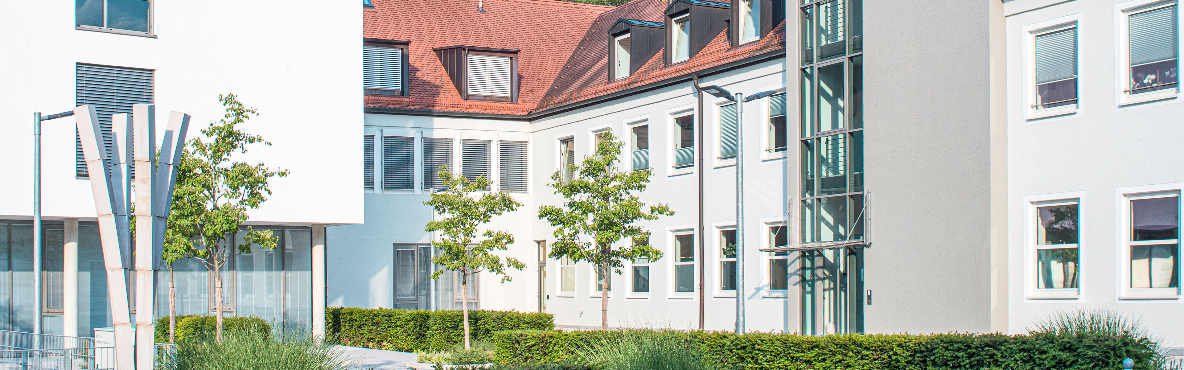 Innenhof mit weißen Fassaden, großen Glasfenstern, roten Ziegeldächern und vereinzelten Bäumen, umgeben von gepflegten Hecken, bei strahlendem Sonnenschein.